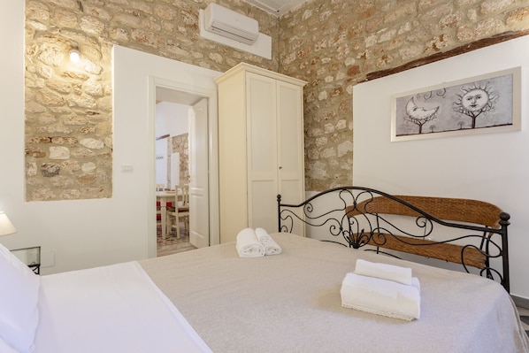 Une chambre avec un mur en pierre, un lit avec une tête de lit en métal, une armoire blanche et un tableau accroché au mur.