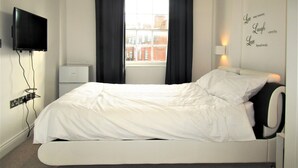 A bedroom with a bed, a flat-screen TV mounted on the wall, a window with black curtains, and a wall with inspirational text.