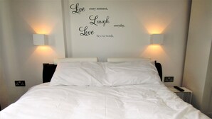 A neatly made bed with white linens, two wall-mounted lamps on either side, and a motivational quote on the wall.
