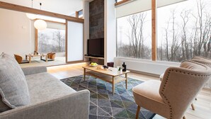 A modern living room with a sofa, armchair, and coffee table. There is a fireplace and a large window with a view of trees.
