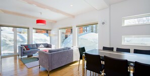 Uma sala de estar moderna com sofá cinzento, uma mesa de jantar com cadeiras, grandes janelas com vista para a neve e uma luminária pendente vermelha.