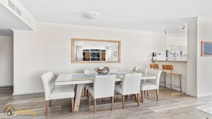 A dining area with a white table and chairs, a wooden-framed mirror, and a kitchen in the background.