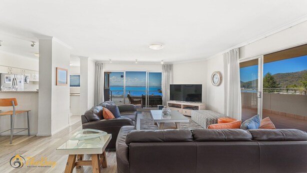 A modern living room with a sofa, coffee table, and TV, a balcony with a view of the sea, and a kitchen area.