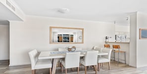 Zona pranzo con tavolo e sedie bianche, uno specchio con cornice in legno e una cucina sullo sfondo.