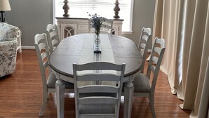 A dining room with a round table, six chairs, two lamps, a vase with flowers, a painting on the wall, and a window with blinds. - Myrtle Beach Townhome on Legends Golf & Resort (Myrtle Beach)