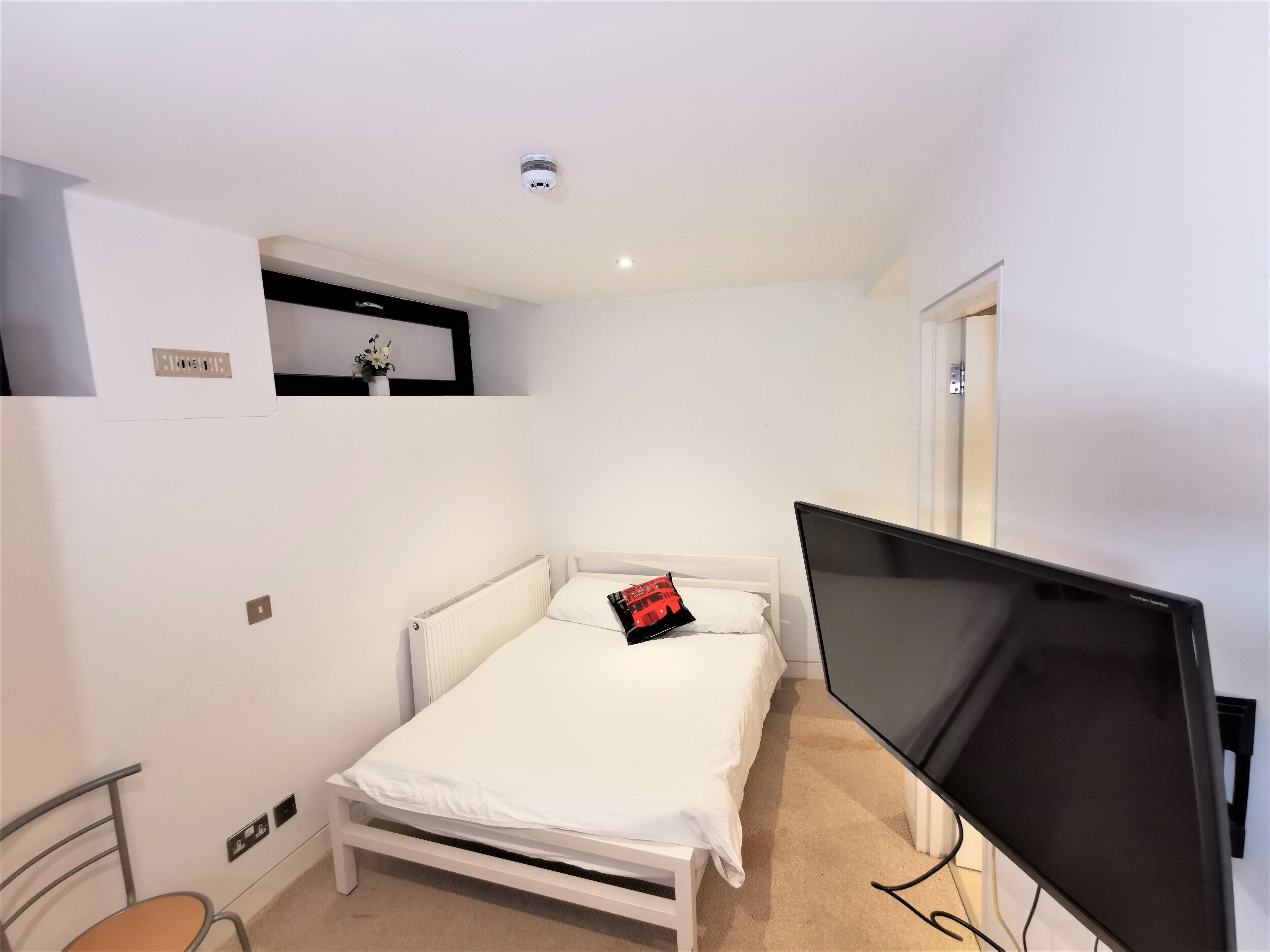 A small, neatly arranged bedroom with a single bed, a television, and a small shelf with a vase.