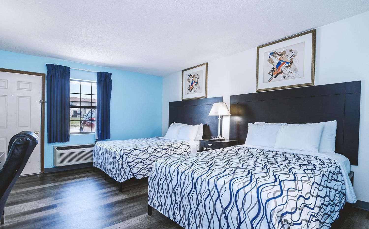 A hotel room with two beds, a desk, a chair, a window with curtains, and artwork on the walls.
