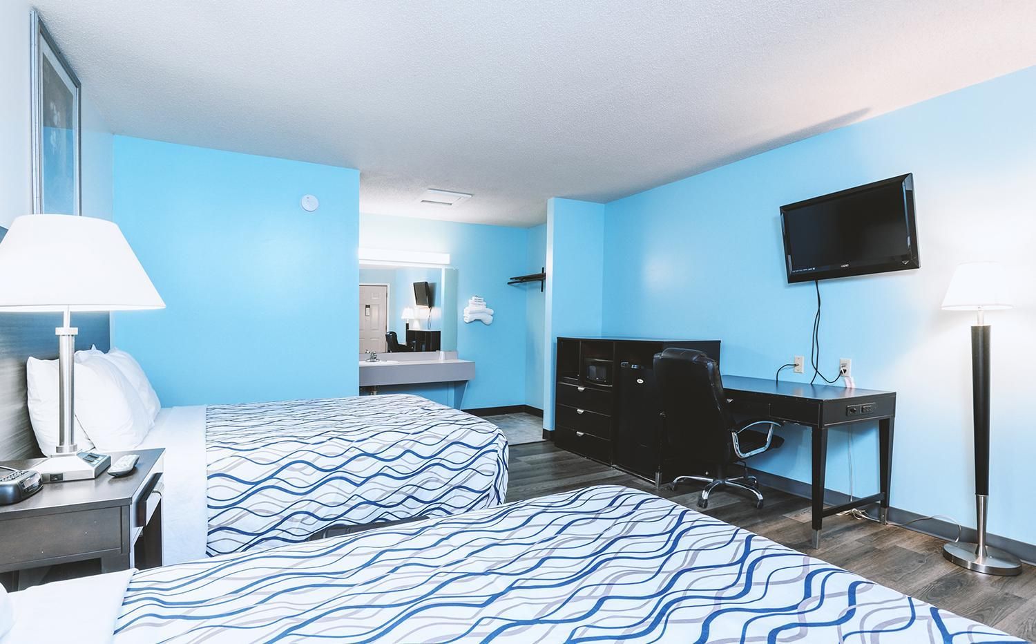 A hotel room with two beds, a desk, a chair, a TV, and a bathroom visible in the background.