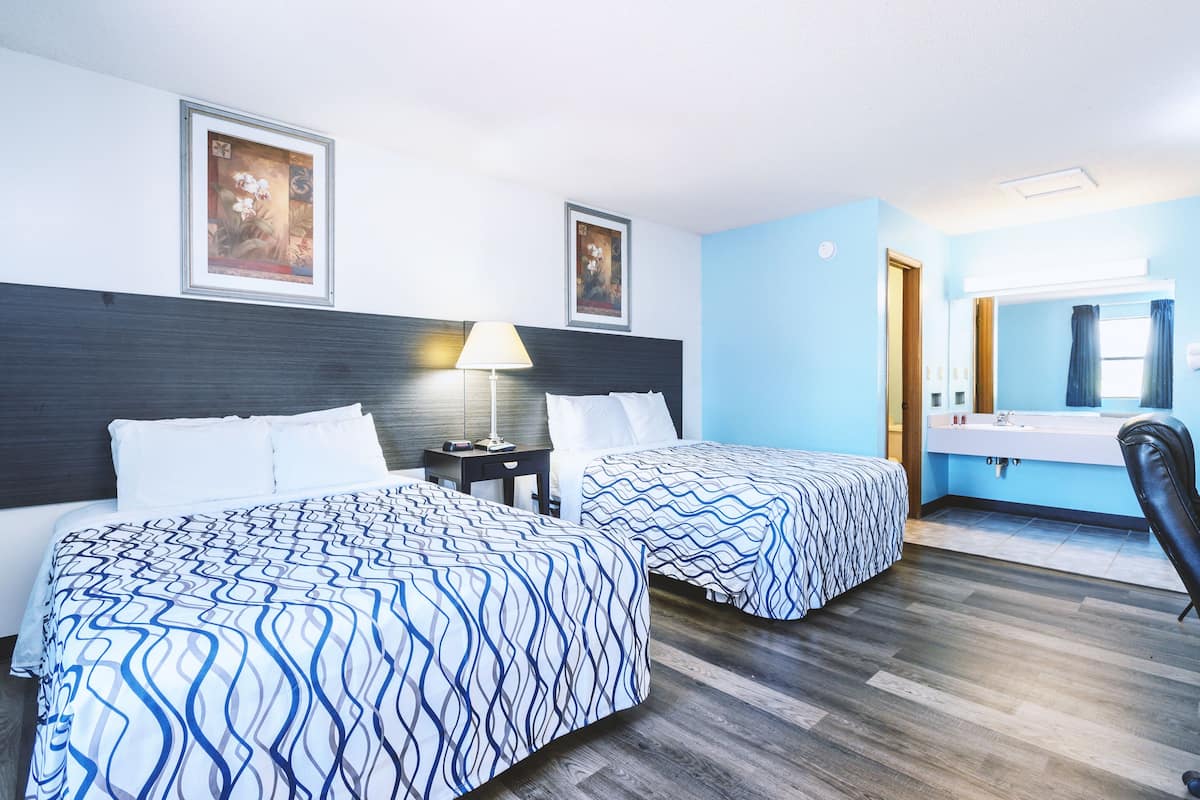 A hotel room with two beds, a nightstand, a lamp, a mirror, and a bathroom with a sink and mirror.