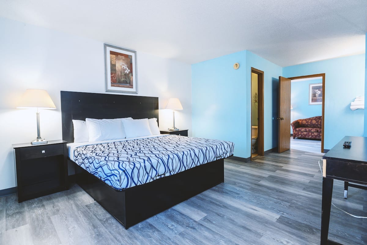 A bedroom with a bed, two bedside tables with lamps, a door leading to another room, and a small table with a remote control.