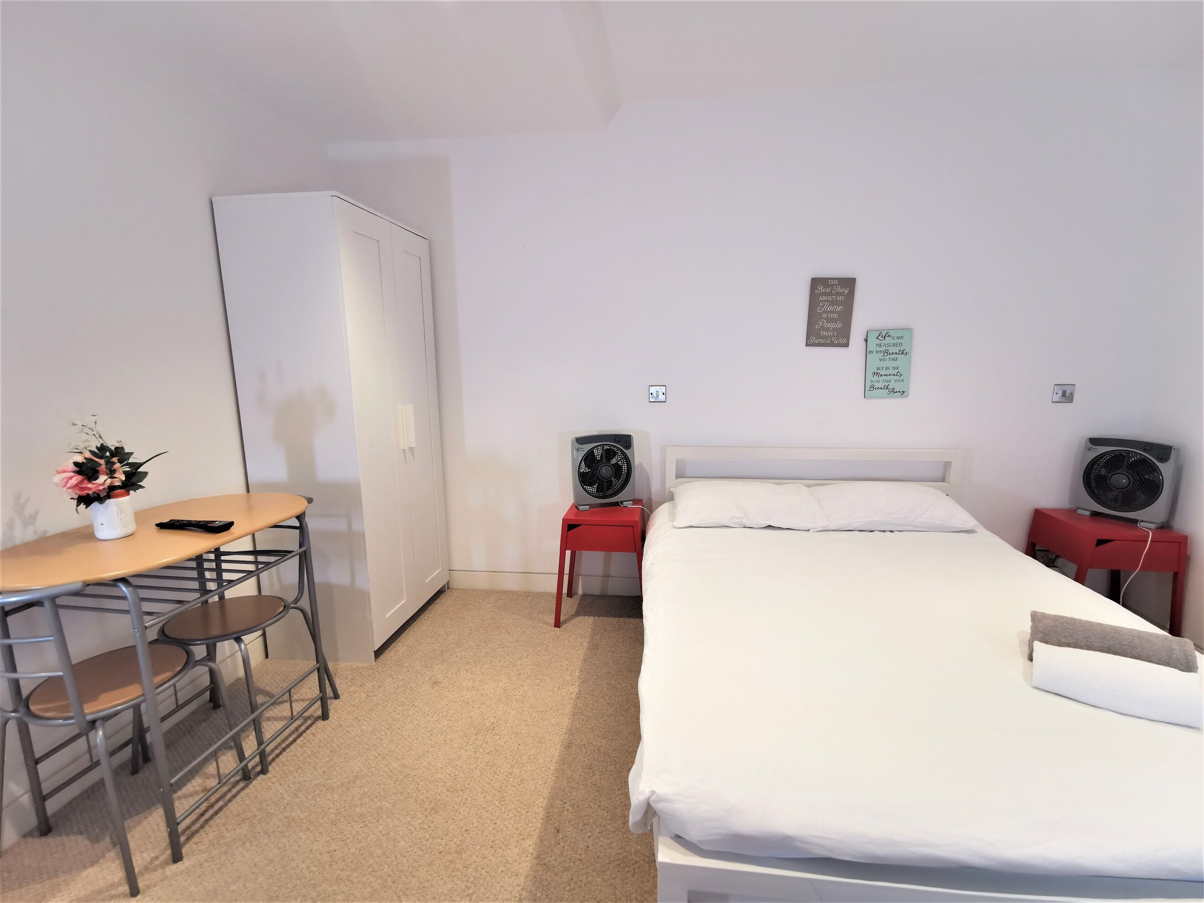 A hotel room with a bed, a desk with a chair, a wardrobe, two bedside tables with fans, and a small table with a vase of flowers.
