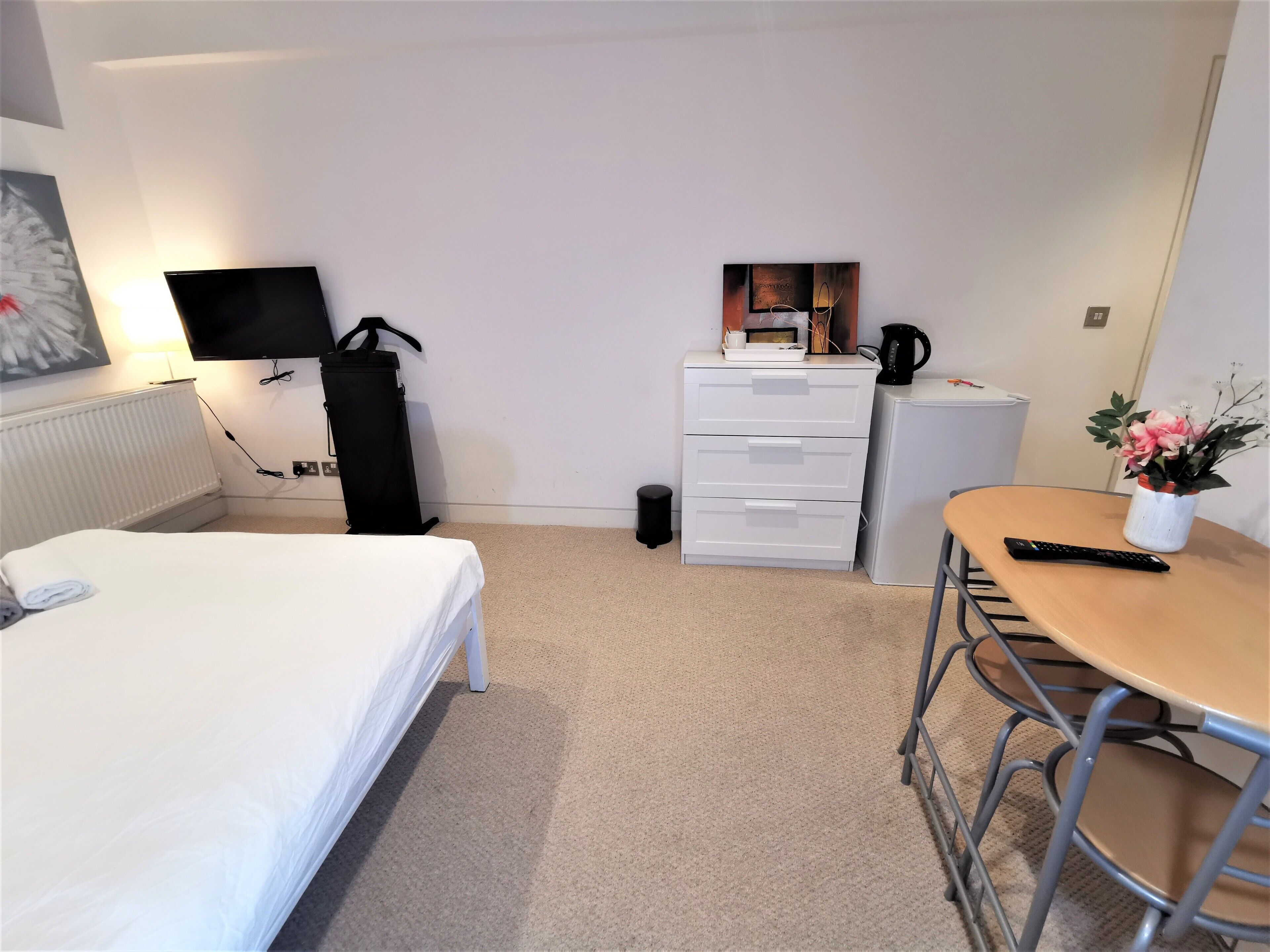 A hotel room with a bed, a desk with a chair, a small refrigerator, a TV mounted on the wall, and a radiator.