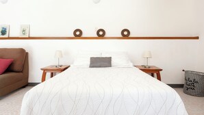 A hotel room with a bed, two bedside tables, a nightstand, a brown sofa, a wall-mounted shelf with decorative items, and a small trash bin.