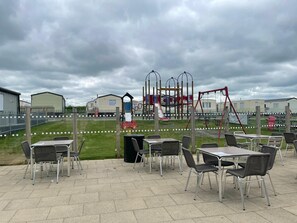 Un espace extérieur aménagé en terrasse avec des tables et des chaises, une aire de jeux avec des toboggans et des balançoires, et un champ d’herbe en arrière-plan.