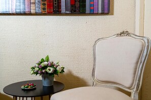 A room with a bookshelf, a chair, a small table with a flower vase, and a plate with food. - Hotel Le Relais De Poste (Arles)