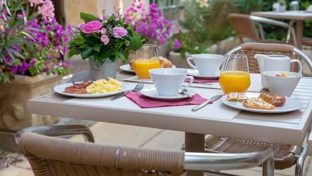 Mesa de desayuno al aire libre con platos de comida, una jarra de jugo de naranja y un jarrón de flores.