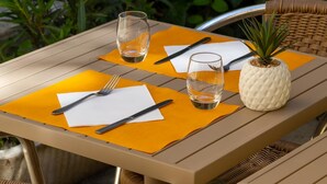 Una mesa al aire libre con una piña como pieza central, dos vasos, cubiertos y servilletas sobre un mantel individual amarillo.