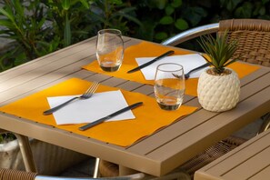 Mesa posta ao ar livre com uma abacaxi como peça central, dois copos, talheres e guardanapos sobre um jogo americano amarelo.