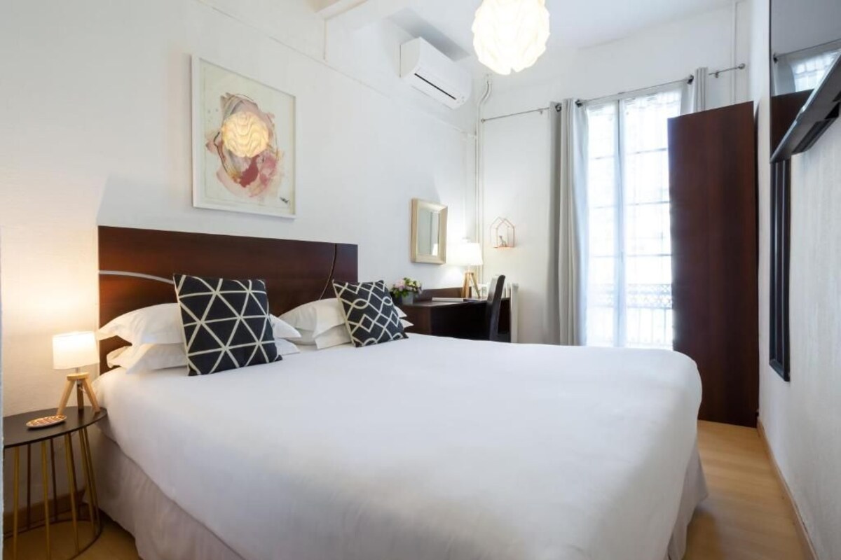 A hotel room with a large bed, a bedside table with a lamp, a desk with a chair, a window with curtains, and a wall-mounted air conditioner.