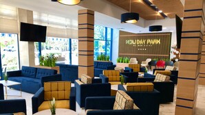 A modern lounge area with sofas and armchairs, a reception desk, and a large window offering a view of greenery. - Holiday Park (Varna)