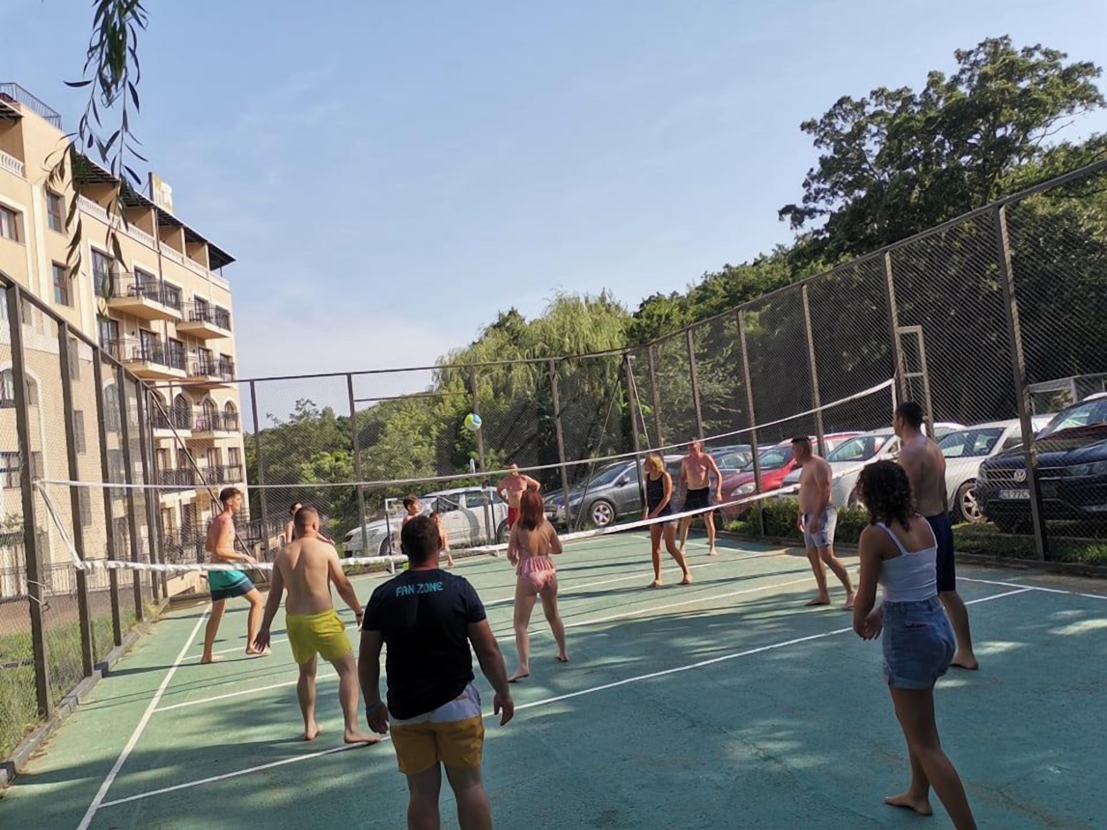 An outdoor tennis court surrounded by a chain-link fence, with a multi-story building in the background.