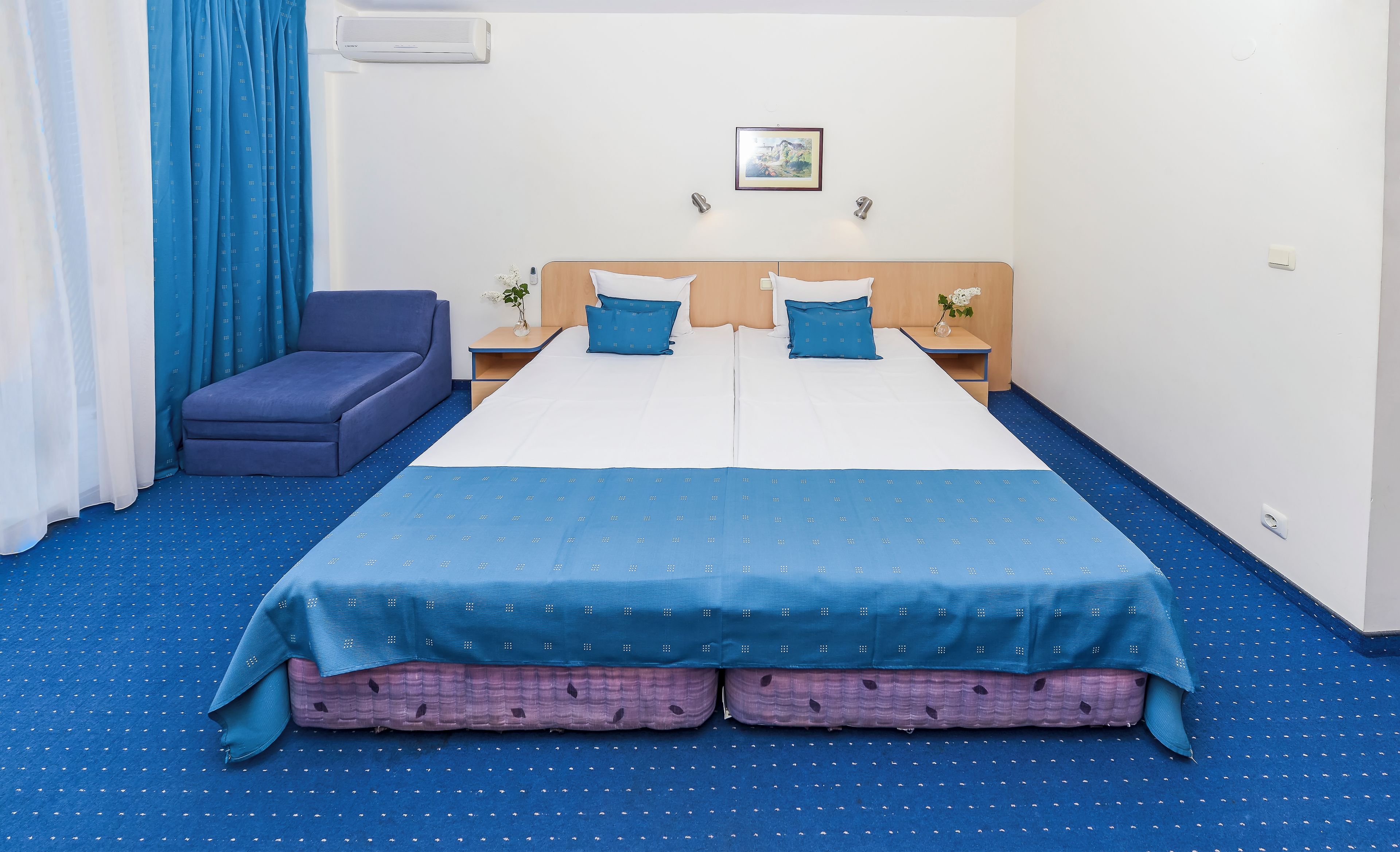 A hotel room with a bed, a sofa, a nightstand, and a wall-mounted air conditioner.
