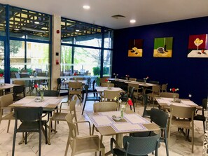 Un restaurant doté de nombreuses tables et chaises, de grandes fenêtres et d’œuvres d’art accrochées aux murs.