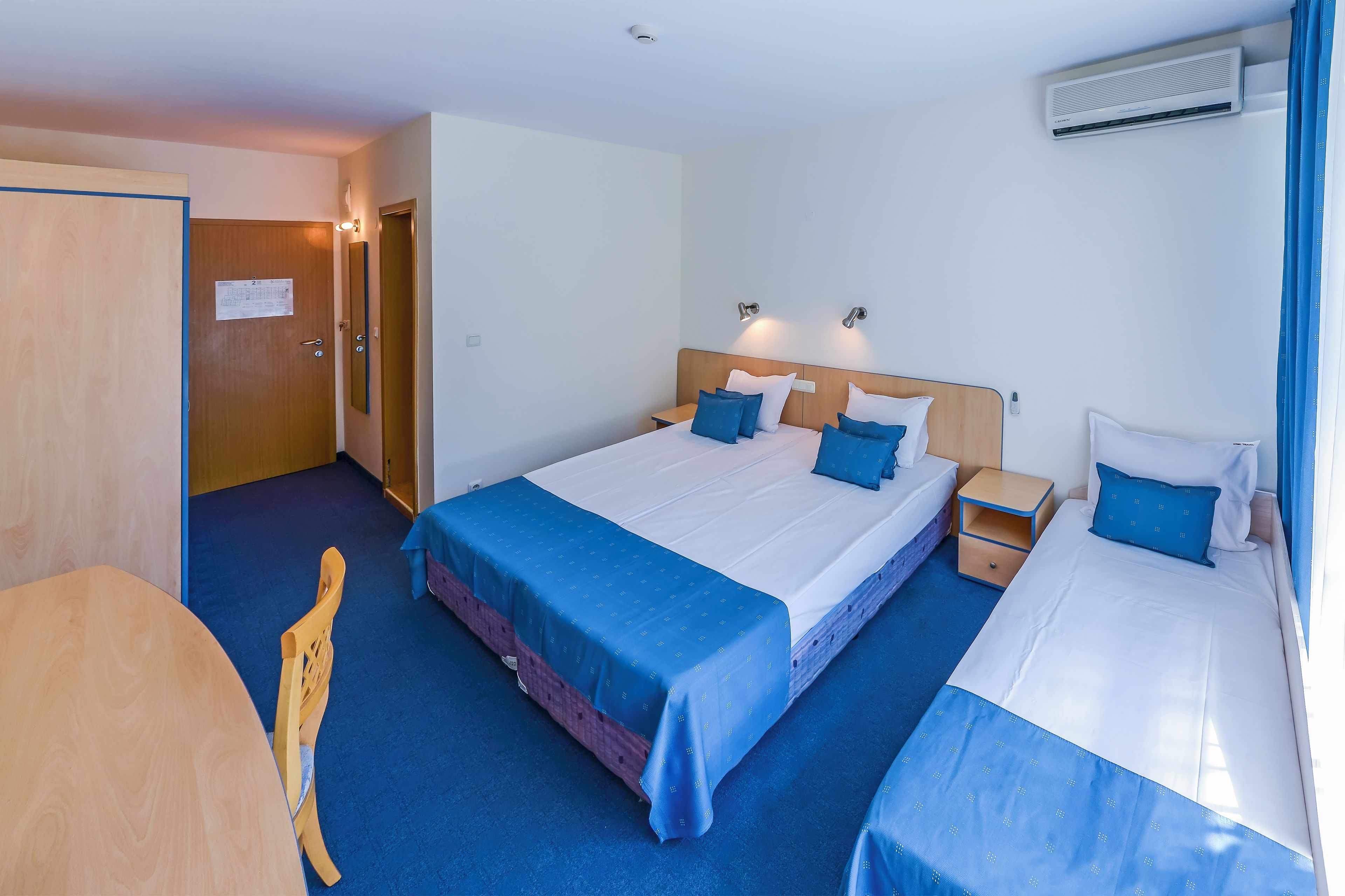 A hotel room with a bed, bedside tables, a chair, a desk, and an air conditioning unit.
