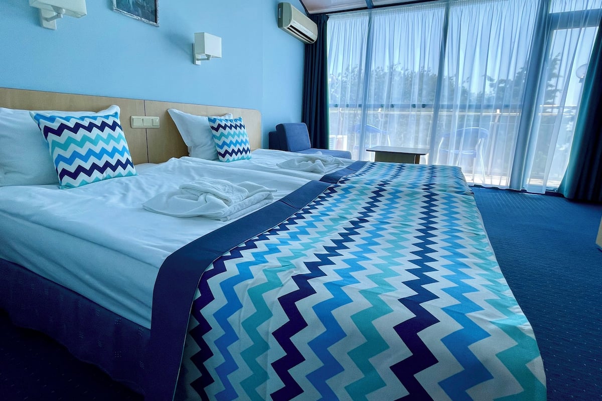 A hotel room with a large bed, a small table, and a view of the outdoors through large windows.