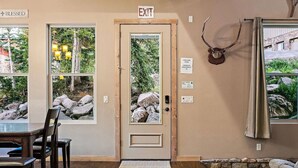 Una stanza con una porta in legno contrassegnata da “EXIT”, un muro in pietra, una testa di cervo appesa e vista su rocce e vegetazione all’esterno.