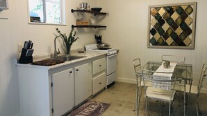 Una cocina compacta con armarios blancos, estufa integrada, lavamanos y comedor con sillas metálicas y una mesa de vidrio.