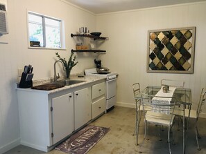 Una cocina compacta con armarios blancos, estufa integrada, lavamanos y comedor con sillas metálicas y una mesa de vidrio.