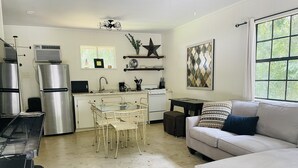 Una cocina compacta con electrodomésticos de acero inoxidable, una zona de comedor con mesa de vidrio y sillas de metal, un sofá blanco con cojines estampados, y un amplio ventanal con vistas a zonas verdes.