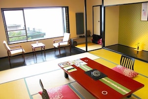 A room with tatami mats, a low table with a clock, and a large window with a city view.