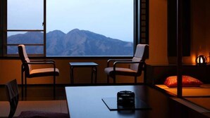 A room with a large window offering a mountain view, two chairs, a small table, and a bed with a red pillow. - Yorokobi no Yado Shinki (Shibukawa)