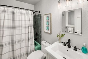 A bathroom with a white toilet, a green bathtub, a white sink with black faucets, a white shower curtain, and a framed picture on the wall.