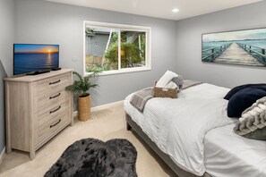 A bedroom with a bed, a dresser, a TV, a window with a view of greenery, and a painting on the wall.