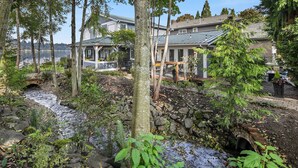 A residential area with a stone bridge over a stream, surrounded by lush greenery and houses with metal roofs.