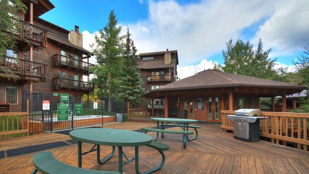 A wooden deck with picnic tables and a barbecue grill, surrounded by multi-story buildings with balconies. - Ski In-Out on 4Oclock with Chair and Town Access! (Breckenridge)