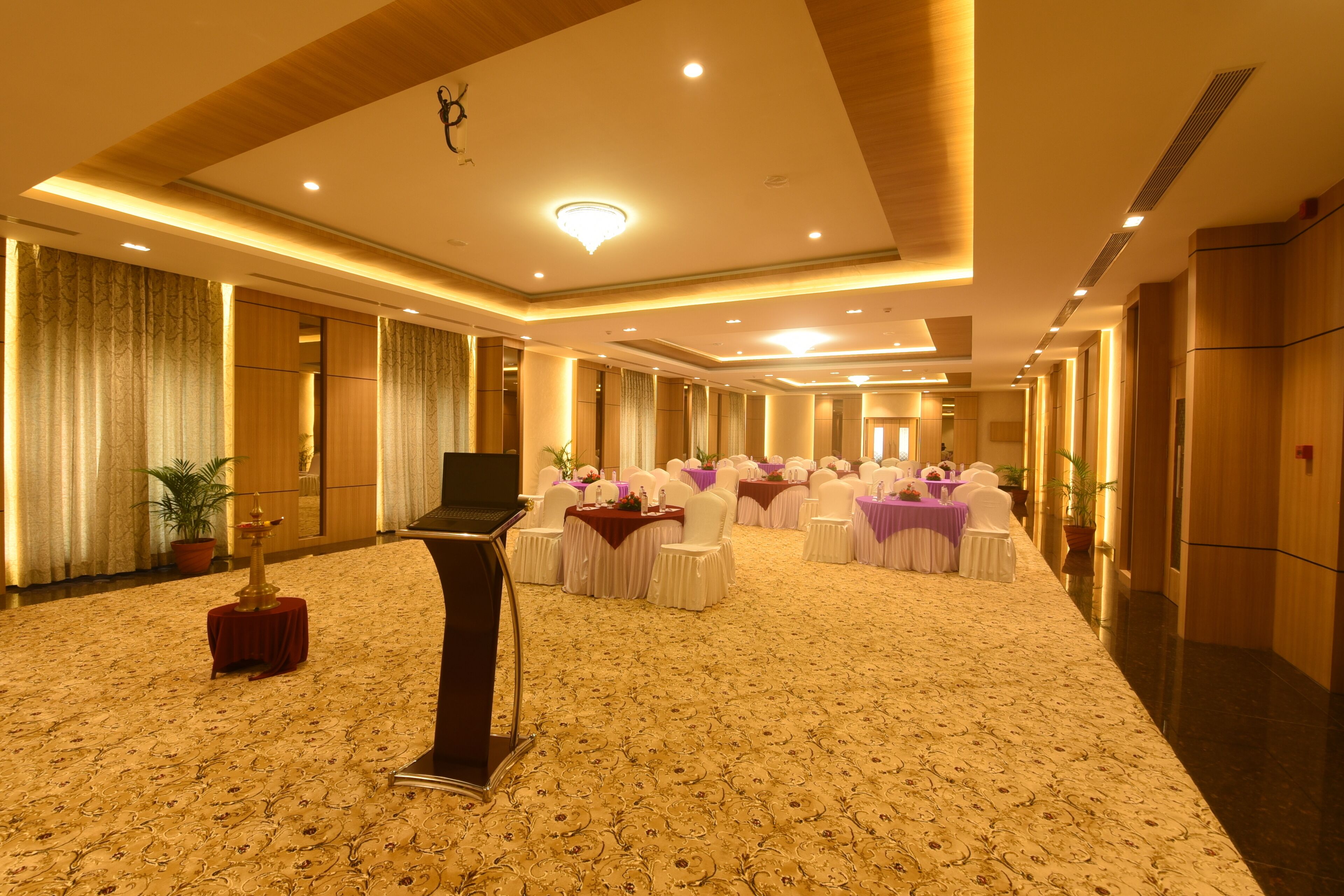 A spacious hotel lobby with a carpeted floor, rows of tables with white chairs, and a stage with a podium.