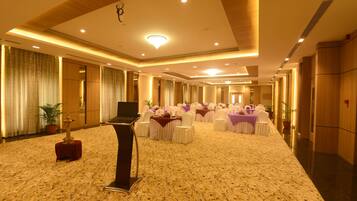 A spacious hotel lobby with a carpeted floor, rows of tables with white chairs, and a stage with a podium.