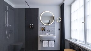 Une salle de bain moderne avec une douche, un lavabo et des toilettes. Il y a deux miroirs, un au-dessus du lavabo et un autre sur le mur. La salle de bain est dotée d’une fenêtre avec des rideaux.