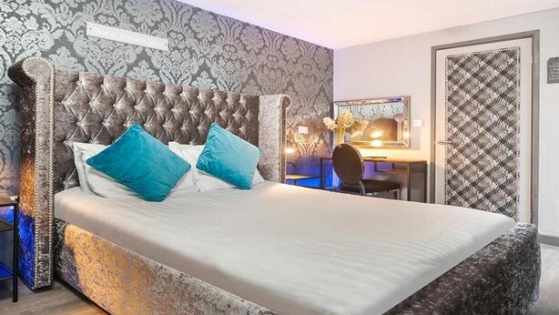 A bedroom with a tufted headboard, a bed with white bedding and two blue pillows, a desk with a chair, and a patterned wall. - OYO Grand Central (Liverpool)