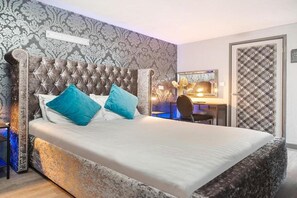 A bedroom with a tufted headboard, a bed with white bedding and two blue pillows, a desk with a chair, and a patterned wall.