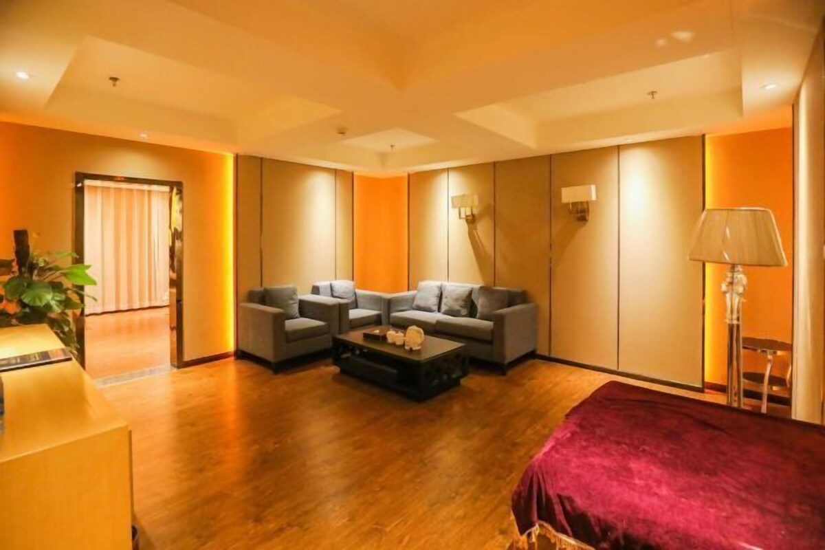 A modern hotel room with a wooden floor, a red bedspread, two grey sofas, a coffee table, and a large window with curtains.