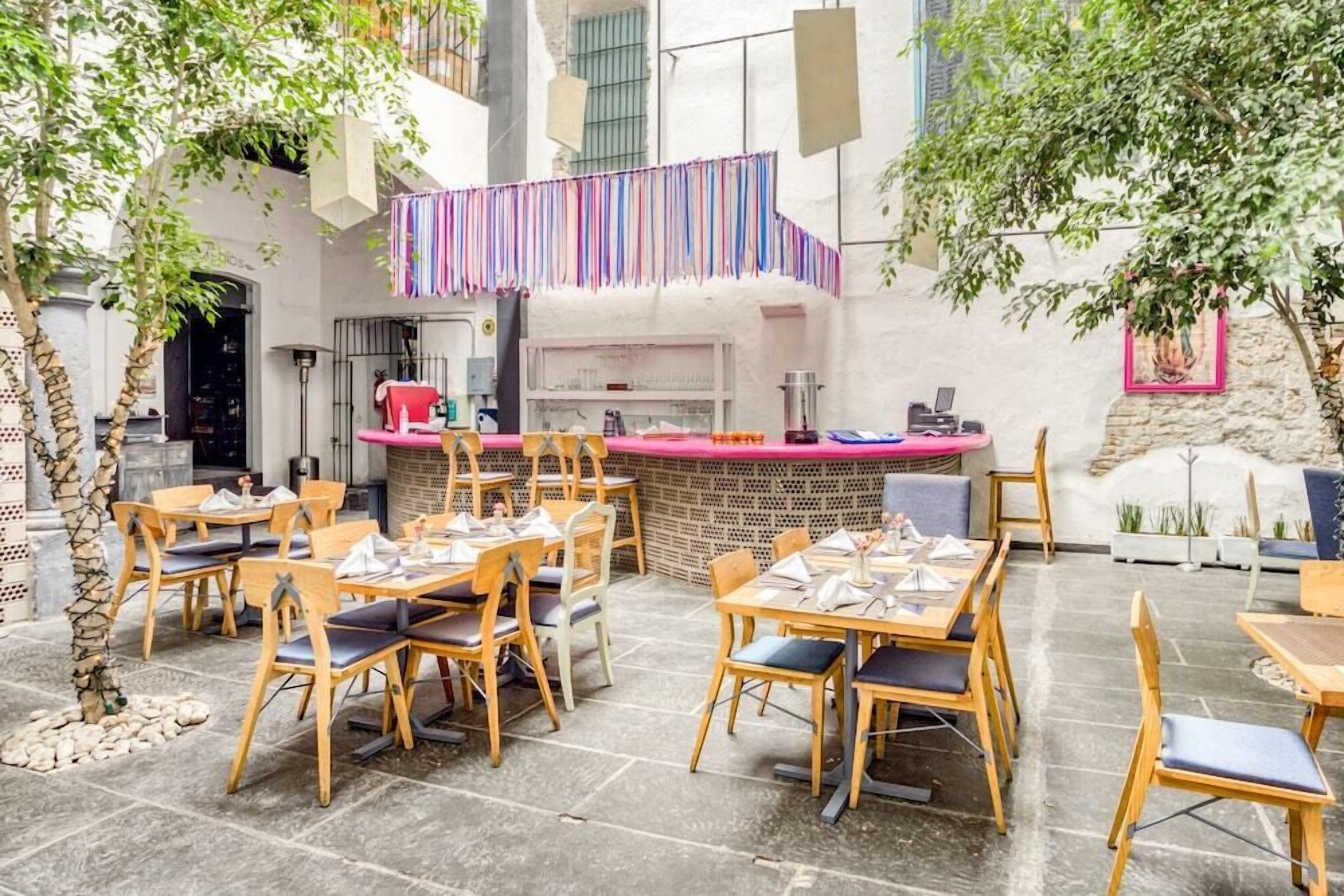 An outdoor dining area with wooden tables and chairs, a bar area with a counter, and a staircase with colorful curtains.