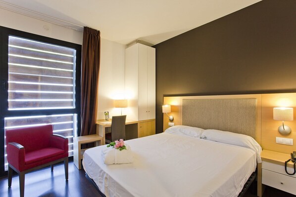 A hotel room with a large bed, a desk with a chair, a red armchair, and a window with blinds.