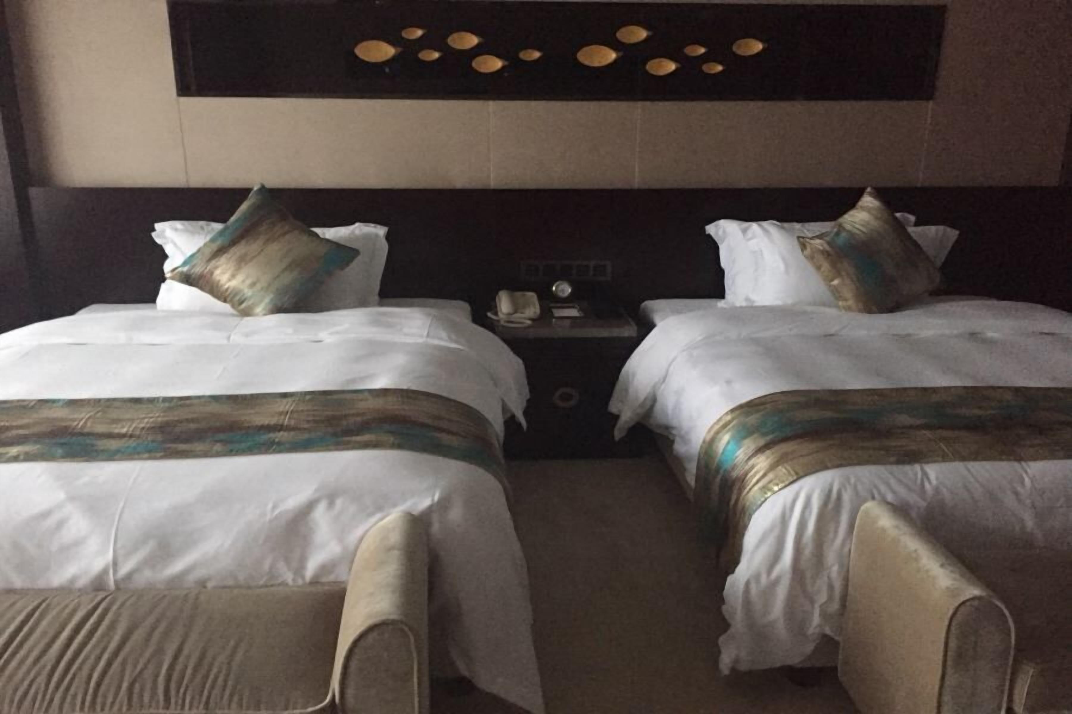 Habitación de hotel con dos camas, cada una con ropa blanca y almohadas decorativas. Se observa un mesita de noche con un teléfono y una pequeña mesa entre las camas. La habitación tiene un cabecero oscuro con adornos circulares.