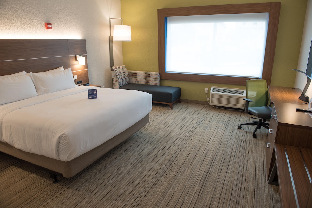 Une chambre d’hôtel avec un grand lit, un bureau avec une chaise, une fenêtre avec des stores, un appareil de climatisation et une lampe de chevet.