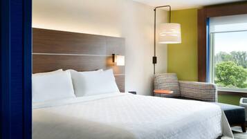 Une chambre d’hôtel avec un grand lit, une table de chevet, une chaise, une lampe et une fenêtre donnant sur des arbres.